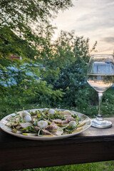 Delicious salad with duck, pear and arugula, and glass of white wine on wooden railings of summer terrace. Soft evening light and lush greenery create romantic atmosphere for outdoor dinner