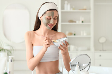 Beautiful young happy woman with mirror and bowl of clay mask at home
