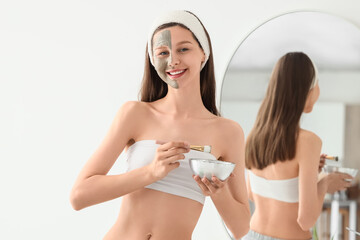 Beautiful young happy woman with clay mask and brush near mirror at home
