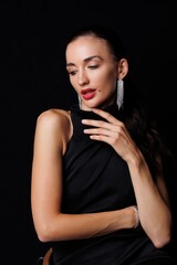 Naklejka premium Beautiful young woman in a black elegant dress posing in studio on black background. Fashion model with long hair, red lips, and stylish earrings. Glamour portrait, studio photoshoot, beauty 