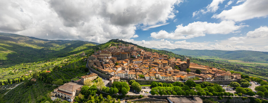 Aerial view of the ancient city atop a hill, terracotta roofs baking under the Tuscan sun, surrounded by lush green hillsides, Cortona, Tuscany, Italy.