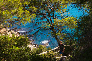 Obraz premium Young boy hiking in nature reserve, during the summer in morning hours, framed between tree branches, in the background we see calm see with turquoise water