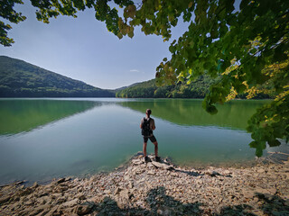 A hiker stands on the rocky lakeshore of Lázbérc Reservoir, admiring the calm water and forested...
