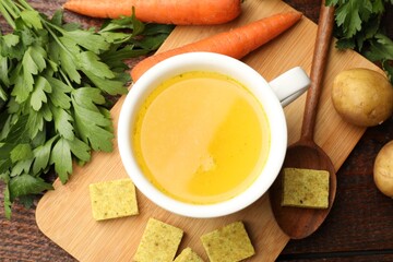 Aromatic bouillon cubes, broth, parsley and vegetables on wooden table, flat lay
