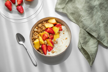 Granola or muesli with natural greek yogurt, strawberry and peach in a bowl on a white table near window
