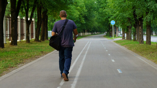 Man walking along a tree-lined pathway carrying a bag in a suburban area during daylight hours