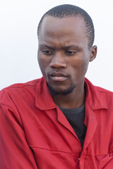 Obraz premium single african construction worker, sited outdoors , in the yard of a warehouse in red workwear uniform standing in front of a warehouse wall