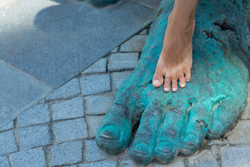 A man's bare foot on top of a bronze sculpture's foot against a backdrop of city cobblestones