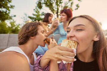 Close-up of young people enjoying burgers and wraps together in park. Concept of social dining, youth lifestyle, street food culture, friendship, and joyful urban moments.