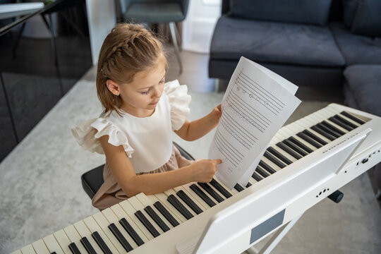 Little girl reading sheet music before piano practice at home. Focused moment of musical learning and preparation in a cozy family setting. - Powered by Adobe