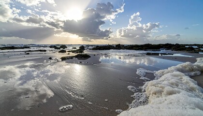 Fototapeta premium Sun shines through clouds reflecting on wet sand and sea foam at the beach