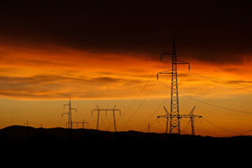 Electricity poles and electric power transmission lines against a vibrant orange sky at sunset on a...