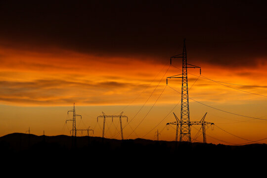 Electricity poles and electric power transmission lines against a vibrant orange sky at sunset on a hot day with flickering air. High Voltage towers provide power supply over a long distance.