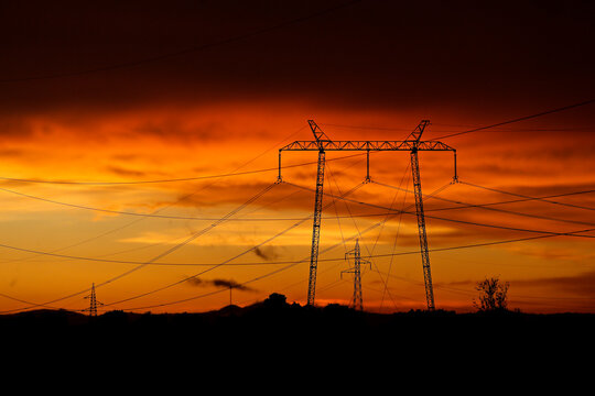 Electricity poles and electric power transmission lines against a vibrant orange sky at sunset on a hot day with flickering air. High Voltage towers provide power supply over a long distance.