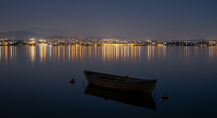A small boat rests on calm water, reflecting city lights at night under a dark blue sky.