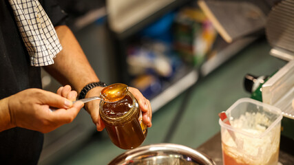 Close-up of person holding a jar of honey with spoon, golden sweet syrup dripping in kitchen scene, concept of natural sweetener and food preparation