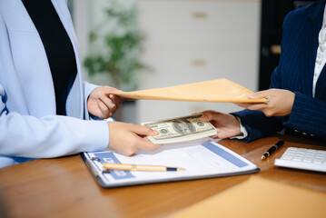 Two businesswomen in formal suits exchange cash over a contract document in an office. Bribery,...