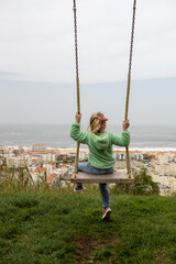 Child swinging high above cityscape, overlooking panoramic ocean view from scenic hilltop playground