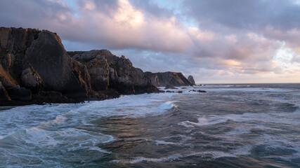 The sun sets over the crashing waves at Praia da Adraga beach in sintra, Portugal, creating a...