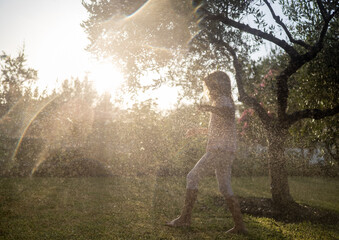 Barefoot kid splashing through water sprinkler, backyard glowing with warm sunset hues, radiating...