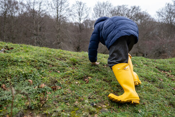 Child crawling up a grassy hill wearing yellow rubber boots and a blue jacket in a park during a...