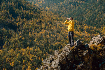 hiker stands on rocky outcrop, enjoying stunning views vibrant autumn foliage and distant mountains under bright blue sky with fluffy clouds. man looks through binoculars in the mountains.