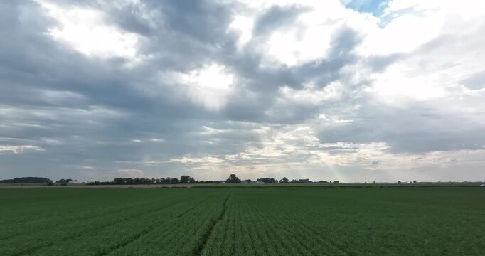 Aerial view of a vast green field stretches towards the horizon under a dramatic sky with sun rays piercing through the clouds, Monroeville, Indiana, United States.