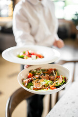 Waiter in white shirt holding two fresh gourmet salads on white plates, featuring greens, cherry tomatoes, watermelon, sesame seeds, and sliced meat.