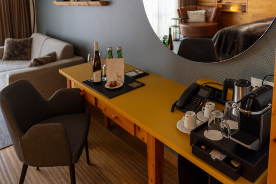 Hotel room with minibar and workspace, wine bottles, sparkling water, coffee machine and welcome treats on yellow wooden desk near large round mirror in cozy stylish interior.