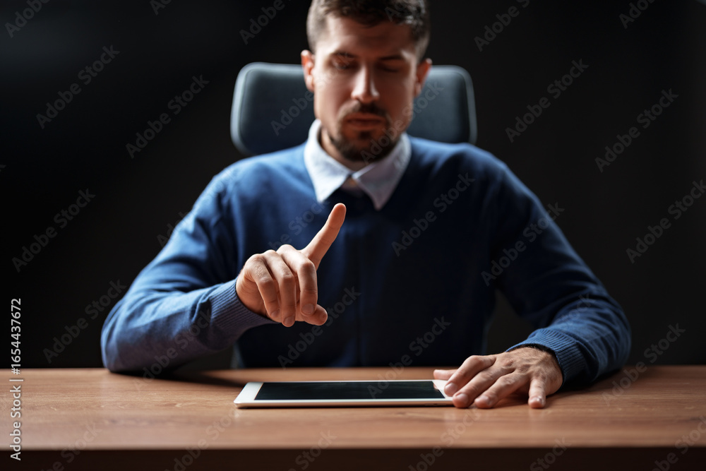 Poster man using tablet while seated at a wooden desk in a dimly lit room during a focused moment of intera