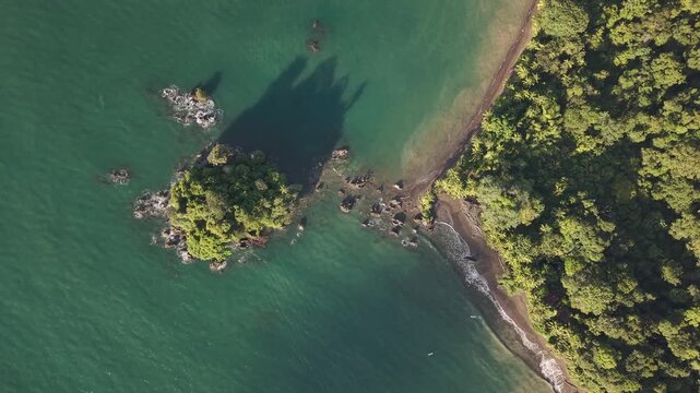 Aerial view of a lush green island casting a long shadow on the turquoise sea near a coastline edged with a dense tropical forest, Nuqu&iacute;, Choco, Colombia.
