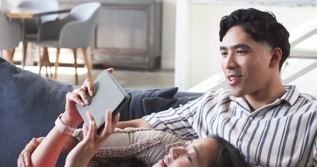 Relaxing diverse couple lying on grey sofa in modern living room, with grey tablet and pillows - Powered by Adobe