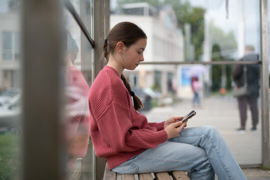 Young girl sitting at bus stop, scrolling through social media feed. - Powered by Adobe