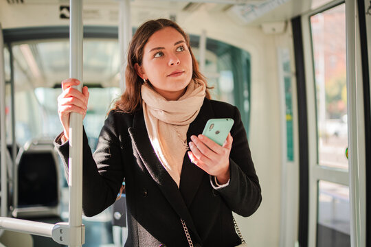 Focused young woman actively using her smartphone while standing in a transportation vehicle, browsing applications with interest, showcasing the daily life of modern commuters navigating their - Powered by Adobe
