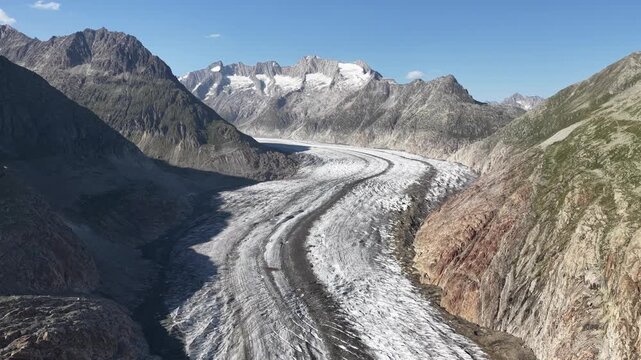 Aerial view of the Aletsch Glacier winding through rocky mountains under a clear blue sky, a striking contrast of ice and stone, Fieschertal, Valais, Switzerland.