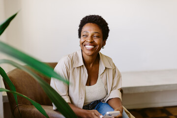 Fototapeta na wymiar Black Brazilian woman smiling and using smartphone on sofa