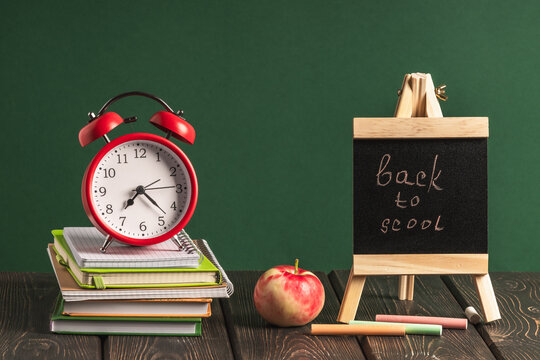 A stack of books and notebooks and a red alarm clock on a wooden table on a green background and a miniature chalkboard with the inscription "back to school".