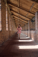 Young woman with long black hair walks through sunlit brick corridor. Walls feature repetitive arch...