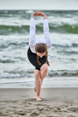 Young woman with brown hair practices yoga on baltic sea beach. Overcast sky and waves create...