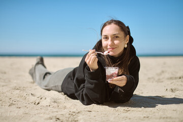 Young woman with brown hair, wearing black hoodie, lies on sandy beach under clear blue sky,...