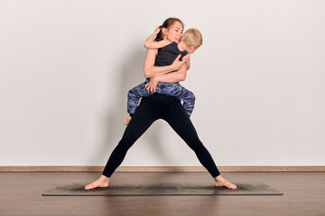 Young woman with brown hair balances small child playfully on yoga mat. Wooden floor and...