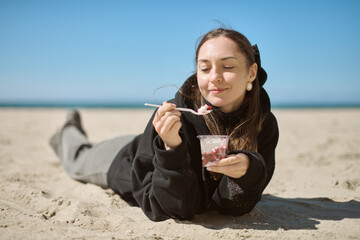 Young woman with brown hair, wearing black hoodie, lies on sandy beach under clear blue sky,...