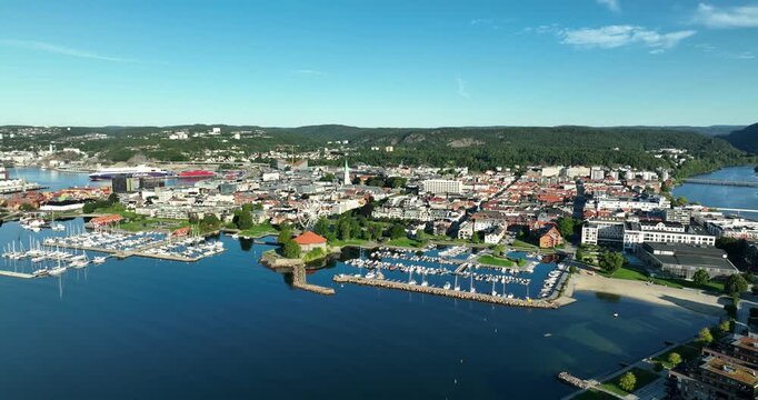 Overlooking a Kristiansand in Norway, this view showcases a marina with boats, surrounded by modern buildings and lush green hills under a clear blue sky, capturing a serene atmosphere.