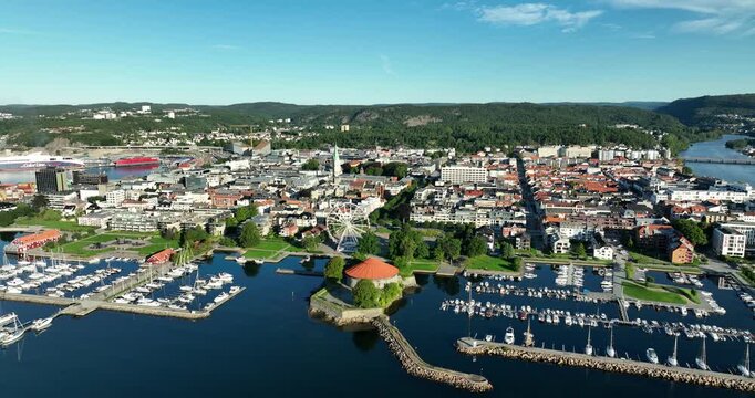 A panoramic view of kristiansand in Norway with numerous boats docked in marinas, vibrant buildings lining the waterfront, and a clear blue sky above.
