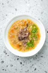 Top view of a bowl of clear broth soup with beef, carrots, celery, and parsley on a speckled gray countertop. Hearty homemade meal with visible ingredients