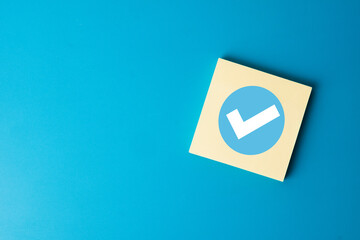 A blue check mark icon on a sticky note. A concept for completion, success, approval, and tasks done. A symbol for productivity, achievement, and finished projects. Blue background with copyspace.