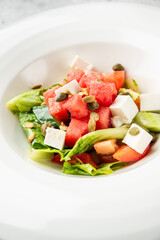 Fresh watermelon salad with feta cheese, lettuce, cucumber, capers, and seeds served in a white...