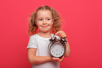 A cheerful young baby girl with curly blonde hair wearing a white shirt is holding a vintage metal alarm clock against a bright pink studio background.