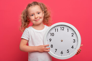 A cheerful young baby girl with curly blonde hair wearing a white shirt is holding a large round wall clock against a bright pink studio background.