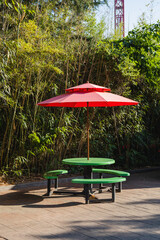 Obraz premium Bright red umbrella and colorful green benches with table for snack in park. Nature background, young bamboo forest. Vertical photo